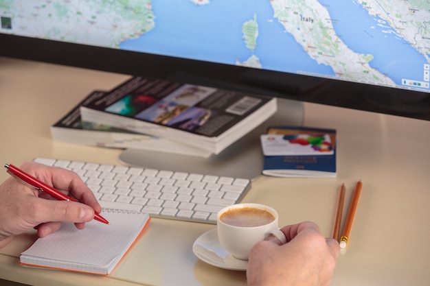 A person using a laptop to compare prices of flights and hotels online. The screen shows various travel websites and the person is taking notes in a notebook. The setting is a cozy home office.