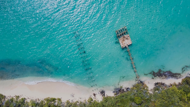 An aerial view of a beach in Southeast Asia with turquoise water and lush green vegetation. There are a few boats anchored in the bay and the sky is clear and sunny.